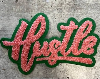 May include: A pink felt patch with the word "Hustle" embroidered in a cursive font on a green felt background.