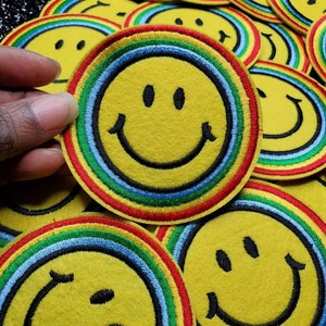 Op de afbeelding: Een close-up van verschillende gele smiley-patches met regenboogkleurige randen. De patches zijn gemaakt van vilt en hebben zwarte geborduurde details.