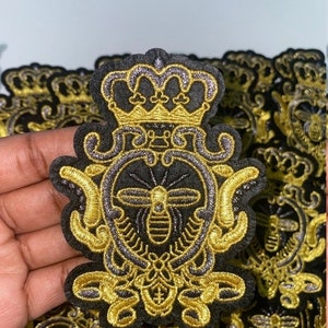 Puede incluir: Parche bordado con una abeja dentro de un escudo con una corona. El parche es negro con bordados dorados y plateados. El diseño es ornamentado y detallado, con una estética regia. Se ven varios parches.