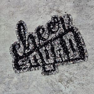May include: A black and silver iron-on patch with the word "Queen" in rhinestones. The patch is shaped like a hand giving the middle finger.