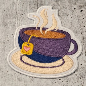 May include: Purple teacup with a yellow tea bag and a heart on it, sitting on a white saucer with steam coming from the cup.