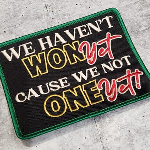 Könnte beinhalten: Schwarzer und grüner gestickter Aufnäher mit weißem und rotem Text, der "We haven't won yet cause we not one yet!" lautet.