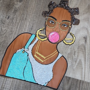 May include: A colorful embroidered patch featuring a woman with brown skin, wearing a white tank top, blue overalls, and large gold hoop earrings. She has a pink bubble in her mouth and is wearing sunglasses. The patch has the words "Melanated" and "BlackQueen" embroidered on it.