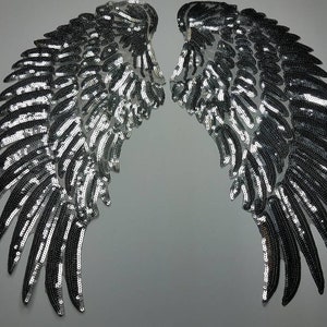 May include: Two silver sequined angel wings. The wings are symmetrical, with each wing featuring a layered design of shimmering sequins. The sequins catch the light, creating a sparkling effect. The wings are likely for costume or craft projects.