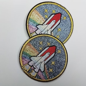 May include: Two round iron-on patches with a space shuttle design. The patches have a blue background with a rainbow glitter effect and white stars. The space shuttles are white with red accents.