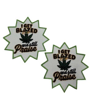 Puede incluir: Dos parches bordados en forma de estrella con centros de tela blanca y bordes cosidos en verde. Cada parche presenta el texto negro I GET BLAZED y and still Praise, con un halo dorado sobre un gráfico de hoja de cannabis verde oscuro.