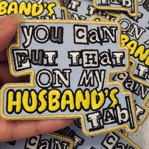 Può includere: Una toppa da stirare ricamata blu e gialla con il testo "You can't put that on my husband's tab".