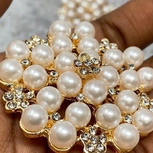 Op de afbeelding: Een broche van goudkleurig metaal met een groot, rond ontwerp met een cluster van witte nepparels en heldere strass.