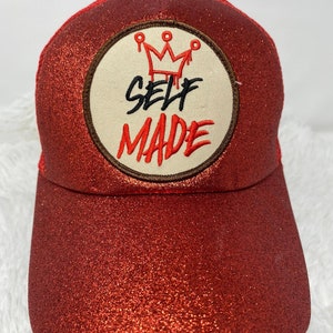Puede incluir: Gorra de camionero roja con purpurina con un parche blanco que dice "SELF MADE" con una corona sobre el texto.