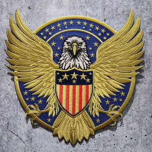 May include: Embroidered patch featuring a golden eagle with outstretched wings, a blue background, and a shield with red and white stripes. The patch has a circular design with stars and is likely for apparel or accessories.