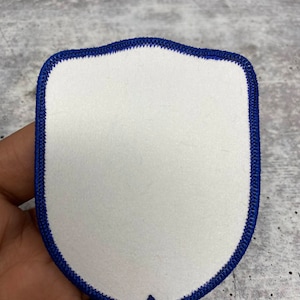 May include: A white, blank, felt patch with a blue stitched border in the shape of a shield.