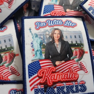 May include: A white fabric patch with a blue border. The patch features a portrait of Kamala Harris, the Statue of Liberty, the White House, and an American flag. The text "I'm With Her" and "Kamala Harris" are printed on the patch.
