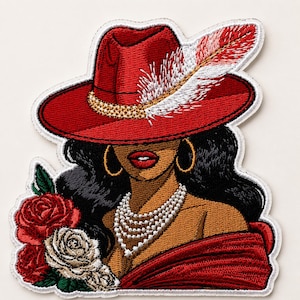 Puede incluir: Parche bordado que representa a una mujer con un sombrero rojo adornado con una pluma, pendientes de aro dorados y un collar de perlas. El diseño incluye rosas rojas y blancas. La mujer tiene cabello oscuro y labios rojos.