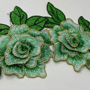 May include: Two green and silver embroidered fabric flowers with gold trim. The flowers are layered and have a 3D effect. The flowers are surrounded by green leaves.