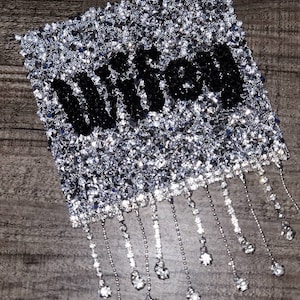 May include: A silver and black rhinestone patch with the word "Wicked" in black rhinestones. The patch has a fringe of silver chains with rhinestones hanging from the bottom.