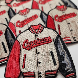 Op de afbeelding: Een wit en rood geborduurde strijk-op patch van een varsity jack met het woord "Capricorn" in rode cursieve letters.