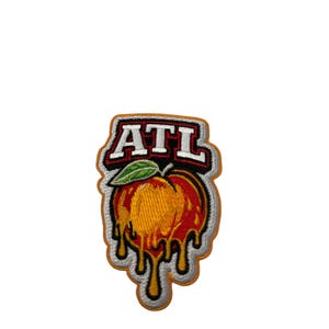 May include: An embroidered patch featuring a peach with orange and red tones, a green leaf, and yellow liquid dripping from the bottom. Above the peach, the letters ATL are stitched in white with a black outline on a red background with a white border.