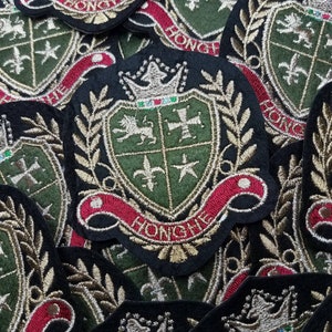 May include: Close-up of several embroidered patches featuring a green and gold heraldic crest with a lion, cross, fleur-de-lis, and star. The crest is surrounded by a wreath of leaves and a red ribbon with the word "HONGKHE" in gold lettering.