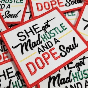 May include: Close-up of several white fabric patches with red, black, and green embroidery. The patches feature the text "She got Mad Hustle and a Dope Soul".