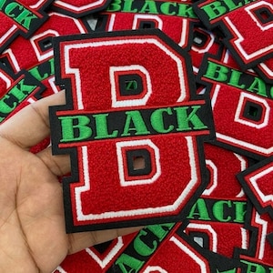 Puede incluir: Parches bordados rojos y negros con la letra "D" y la palabra "BLACK" en verde. Los parches tienen un borde blanco. La imagen muestra varios parches, probablemente para ropa o accesorios.