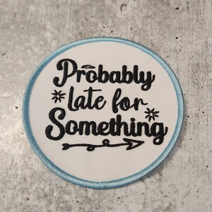 May include: White round patch with a blue border. The patch has black embroidered text that reads "Probably *late for* Something" with a small arrow pointing to the right.