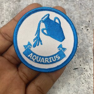May include: A blue and white embroidered iron-on patch with the astrological sign Aquarius and the word "Aquarius" in a banner.