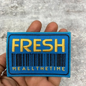 Peut inclure: Un patch brodé bleu et noir avec le mot "FRESH" en lettres jaunes et un code-barres en dessous. Le texte "ME ALL THE TIME" est sous le code-barres.