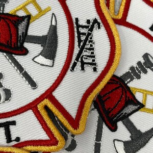 Op de afbeelding: Close-up van een witte brandweerbadge met rode en gouden rand. De badge toont een gekruiste bijl en ladder, een brandweerhelm en een slang.