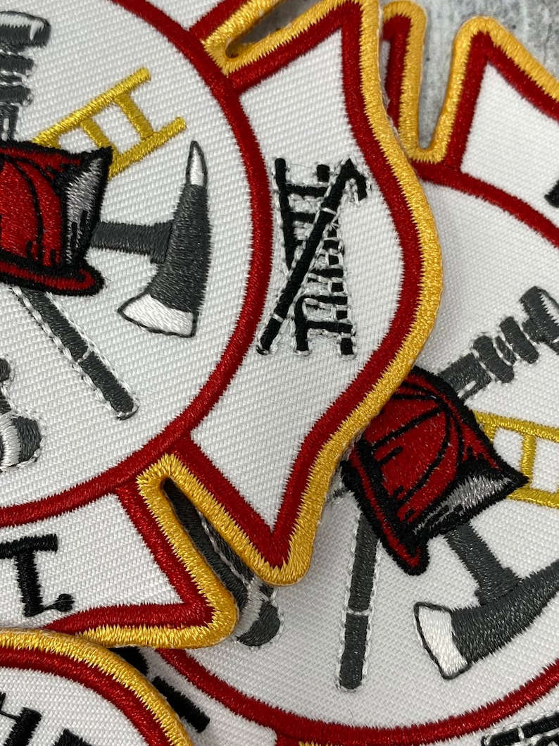 Op de afbeelding: Close-up van een witte brandweerbadge met rode en gouden rand. De badge toont een gekruiste bijl en ladder, een brandweerhelm en een slang.