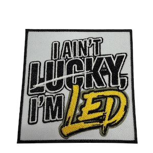 May include: A square embroidered patch with a white background and a thick black border. The text reads I AIN'T LUCKY, I'M LED in bold black lettering, with the word LED featured in a textured, bright yellow thread with a distressed style.