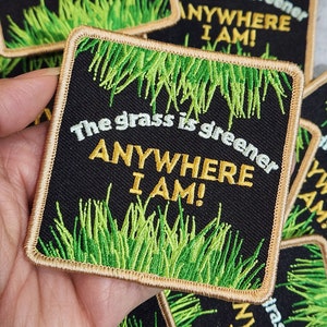 May include: A black and gold embroidered patch with green grass and the text "The grass is greener ANYWHERE I AM!"