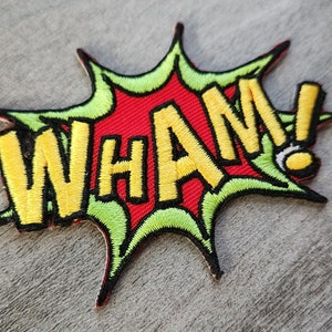 Puede incluir: Un parche bordado amarillo y rojo con la palabra "WHAM!" en una fuente de estilo cómic. El parche está rodeado por un borde verde y tiene forma de estrella.