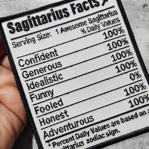 May include: White fabric patch with black text that reads "Sagittarius Facts" and lists traits with percentages. The traits include "Confident", "Generous", "Idealistic", "Funny", "Fooled", "Honest", and "Adventurous".
