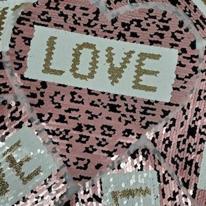 May include: Pink and black sequin fabric with the word "LOVE" written in gold glitter on a white background. The fabric is in the shape of a heart.