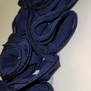 May include: Close-up of a navy blue fabric flower with ruffled edges and black stitching. The flower is made of layered fabric petals, creating a textured, three-dimensional effect. The design is likely for a hair accessory or clothing embellishment.
