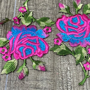 May include: Two embroidered floral patches featuring vibrant pink and blue roses with green leaves and small pink flowers. The patches are set against a wooden background.