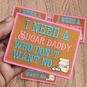 Op de afbeelding: Geborduurde patch met de tekst "I NEED A SUGAR DADDY WHO DON'T WANT NO" in teal en roze op een bruine achtergrond, met een roze rand. Een kleine suikerzak en suikerklontjes zijn ook afgebeeld.
