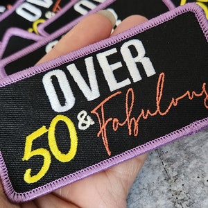 May include: A black fabric patch with purple stitching. The patch says "OVER 50 & Fabulous" in white, yellow, and orange lettering.