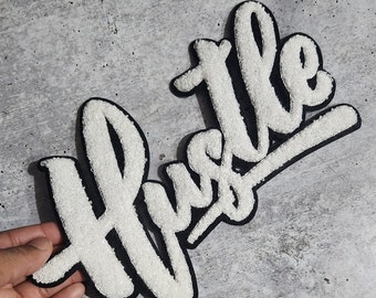 May include: White, textured "Hustle" and "the" patches with black borders. The patches are in a cursive font and are displayed on a grey surface.