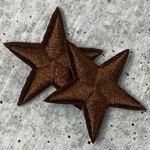 Puede incluir: Dos parches bordados con forma de estrella marrón. Cada estrella tiene cinco puntas y está hecha con un hilo texturizado. Los parches son de color marrón cálido y están colocados sobre un fondo gris.