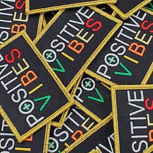 May include: A close-up of a pile of black fabric patches with gold stitching. The patches have the words "POSITIVE VIBES" embroidered on them in white, green, orange, and red thread.
