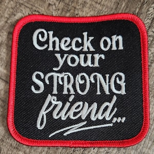 May include: Black square patch with red stitching around the edges. The patch has white embroidered text that reads "Check on your STRONG friend..."
