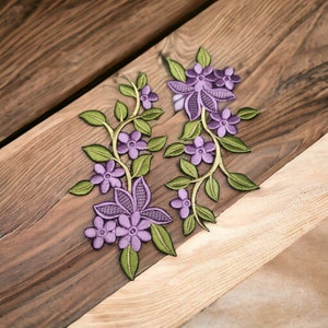 Puede incluir: Dos apliques bordados de flores moradas y verdes con un diseño de vid. Los apliques están sobre una superficie de madera.