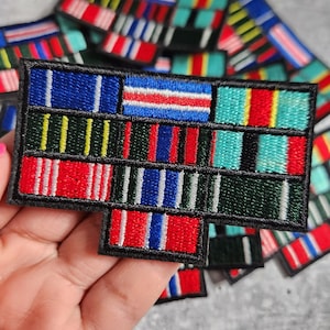 Op de afbeelding: Een zwarte geborduurde patch met een verscheidenheid aan kleurrijke linten. De linten zijn in rijen gerangschikt en zijn van verschillende breedtes en kleuren, waaronder rood, wit, blauw, groen en geel.