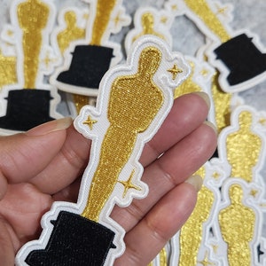 Puede incluir: Un conjunto de parches bordados de color dorado con forma de la estatuilla del Premio de la Academia. Los parches tienen una base negra y están rodeados por un contorno blanco. Cada parche presenta una pequeña estrella brillante.