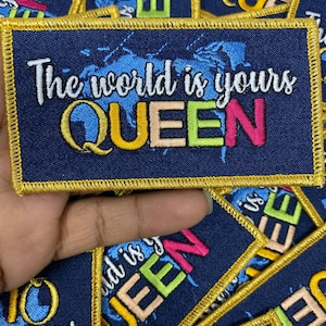 May include: A blue fabric patch with gold stitching around the edges. The patch has a white world map design and the text "The world is yours QUEEN" in colorful embroidery.
