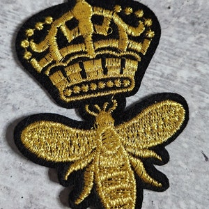 May include: A gold embroidered bee with a black outline and a gold embroidered crown on top. The bee and crown are on a black background.