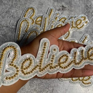 May include: A white fabric patch with the word "Believe" spelled out in gold and silver sequins. The patch is surrounded by other patches with the same word.