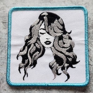 May include: Embroidered patch featuring a black and white illustration of a woman with long, flowing hair. The patch has a turquoise border.
