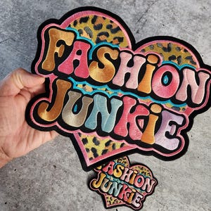 May include: Two embroidered iron-on patches in the shape of a heart with the text "Fashion Junkie" in a colorful retro font. The larger patch has a leopard print background.